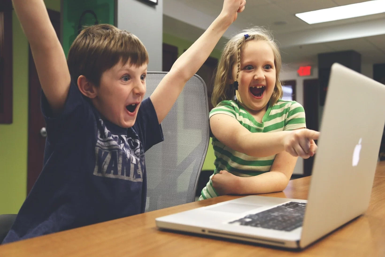 The image shows two happy children, one boy and a girl, and the girl is pointing towards the laptop.